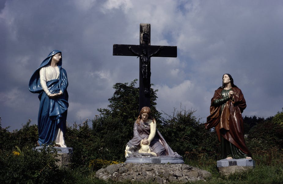 A statue of Saint Brigid seated below a large cross with a statue of a female figure standing on either side.