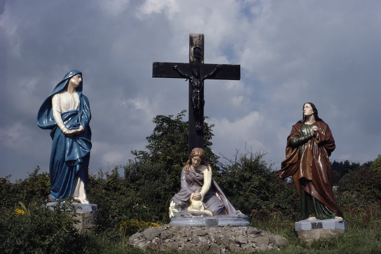 A statue of Saint Brigid seated below a large cross with a statue of a female figure standing on either side.