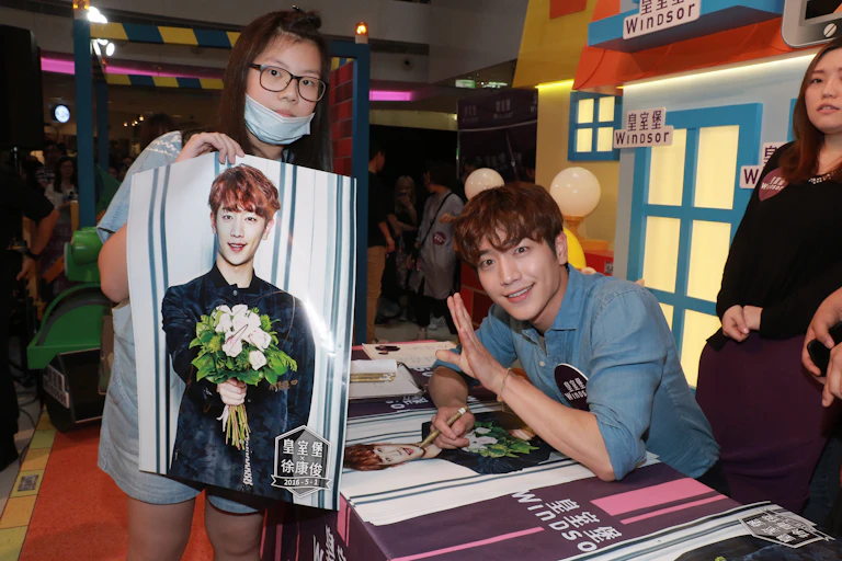 Man holding pen sits at a table next to a woman holding a poster bearing his photo.
