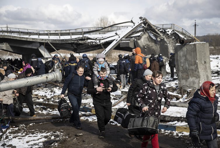 people dressed in winter coats carry their belongings through the snow, with a destroyed bridge in the background
