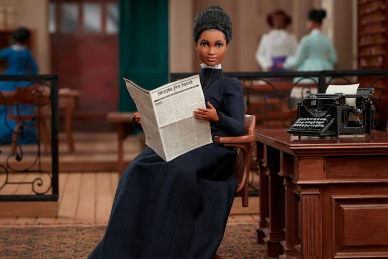 A Black doll in a floor-length 19th-century dress sits in an office reading a newspaper.