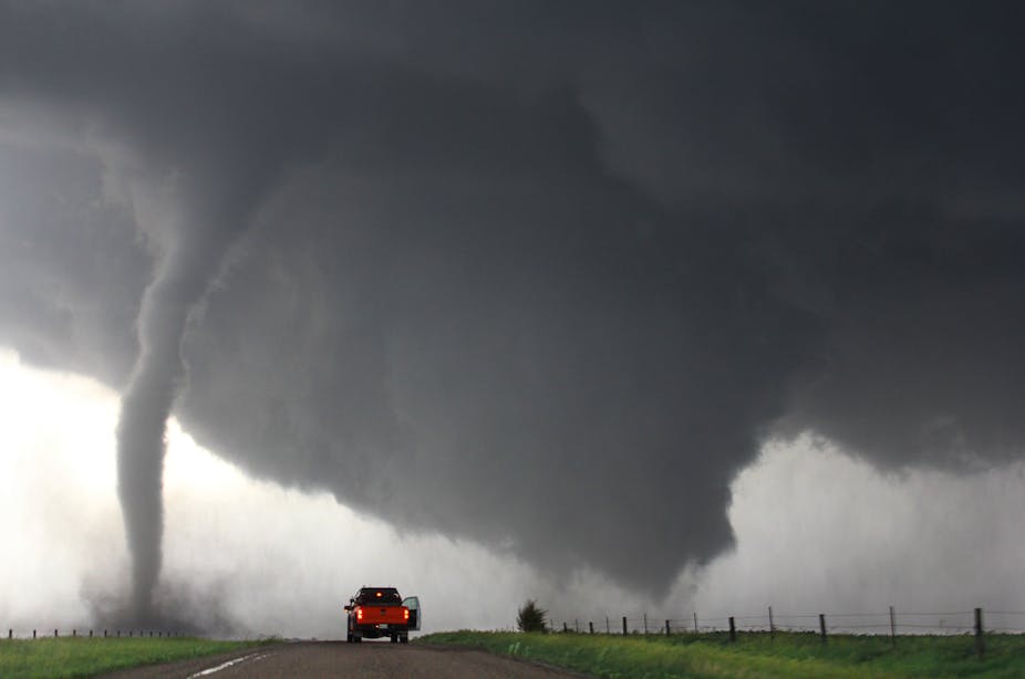 A truck parked on a road with two types of tornadoes on either side, one thin and touching the ground and the other wider.