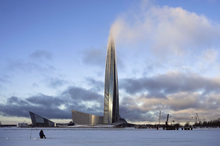 A man fishing on the ice in front of a tall modern office tower.