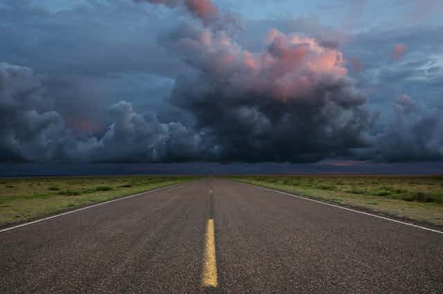 Asphalt road with yellow lines with grass on either side, all running ahead into the distance under a darkening sky of black, blue and orange clouds.
