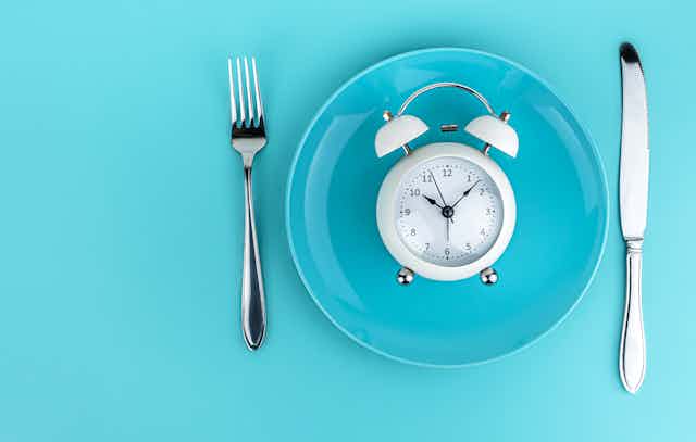A white alarm clock on a blue plate with a fork and knife on either side, on a blue background.