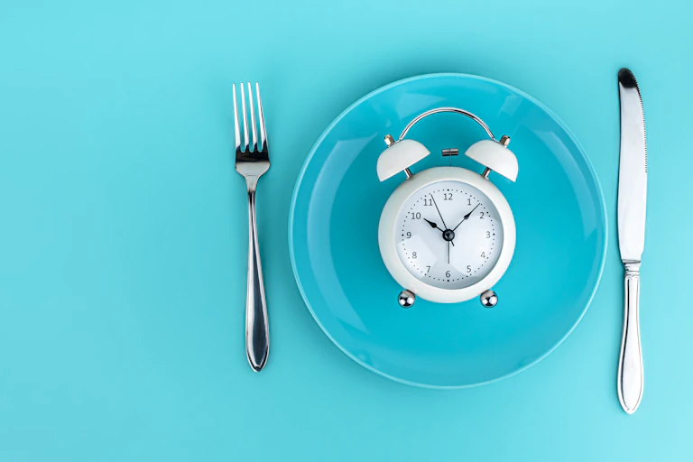 A white alarm clock on a blue plate with a fork and knife on either side, on a blue background.