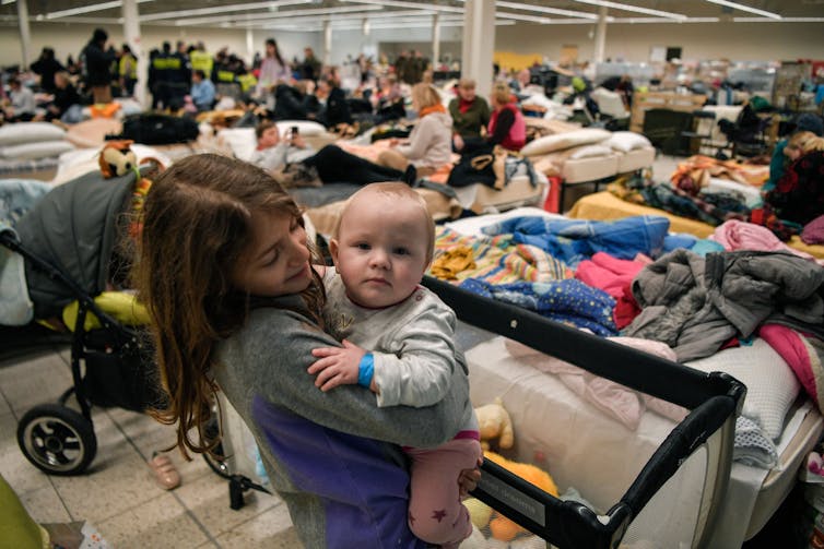 A child holds a baby, looking straight at a camera, with the backdrop of a full room and beds accommodating Ukrainian refugees
