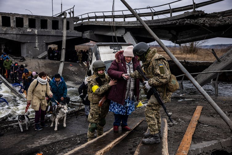 Two soldiers support an elderly woman, wearing a maroon coat and white hat, as she walks over a destroyed bridge in Ukraine. Behind her, a row of people, some of them walking dogs, wait to also cross the remnants of the bridge.