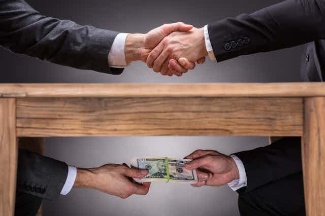 Two people shaking hands on top of a table while they exchange money below the table
