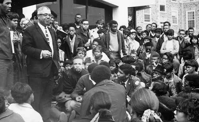 An HBCU president stands among a crowd of Black students who are sitting and standing outside.