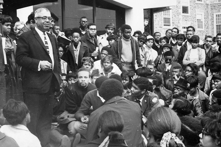 An HBCU president stands among a crowd of Black students who are sitting and standing outside.