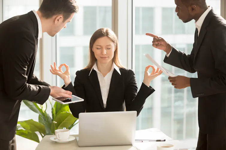 A woman in a suit suits behind a laptop with her arms up in a meditative position. On either side of her, two men in suits are gesturing forcefully while speaking to her.