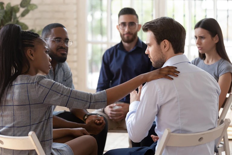 A group of people sitting in a circle. A woman has her hand on a man's shoulder as he gestures to the group.
