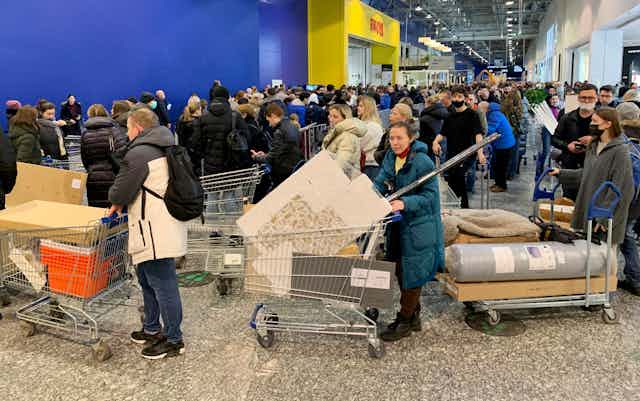 Men and women in coats push carts full of furniture and other products amid a crowd of people outside a blue and yellow Ikea facade