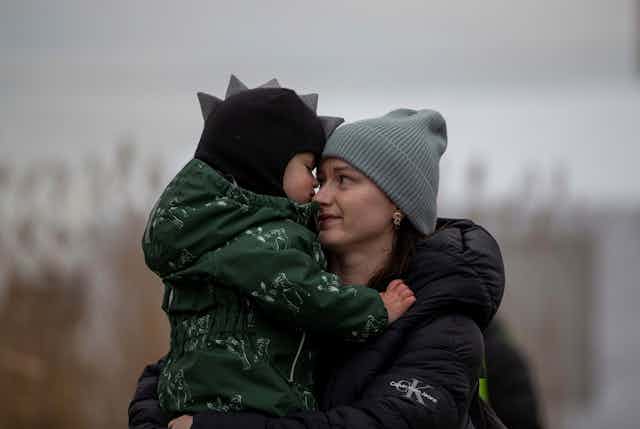 A woman wearing a grey knitted hat holds a small child in a green coat and hat with stegosaurus spines