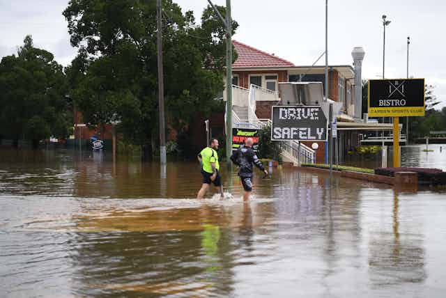 two men walk through floodwaters near sign reading "drive safely"