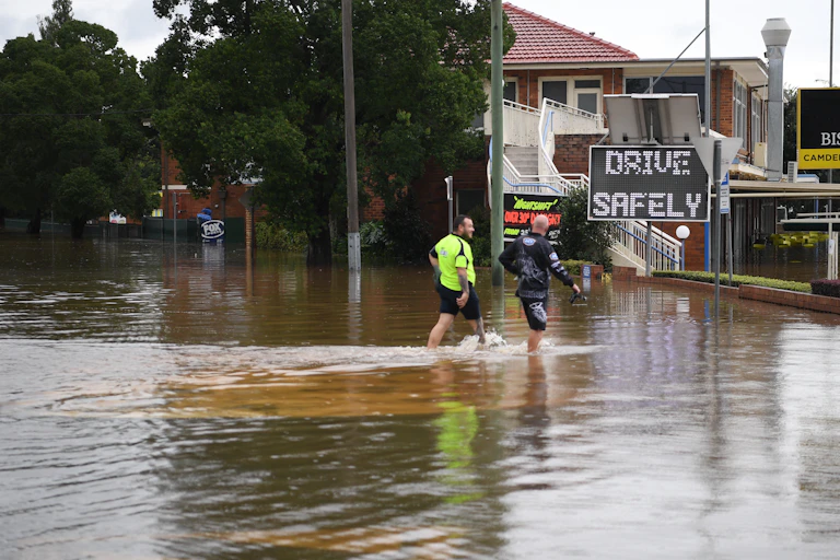 two men walk through floodwaters near sign reading "drive safely"