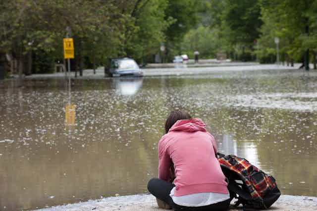 Woman sitting at edge of flood with submerged car in distance
