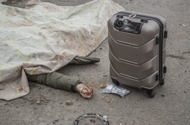A human corpse lies under a blanket with an arm outstretched and an undamaged suitcase nearby.