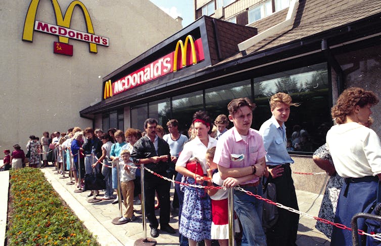 People stand outside a restaurant-looking building with yellow arches spelling an M as they wait to eat McDonalds for the first time.