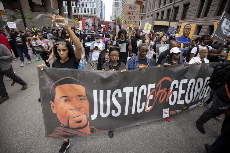 a crowd marches in a city street behind a banner that reads justice for George