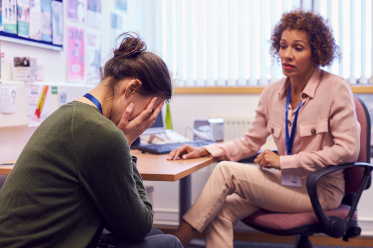 AA seated young woman buries her face in her hands as she sits across from a woman in a chair at a desk.