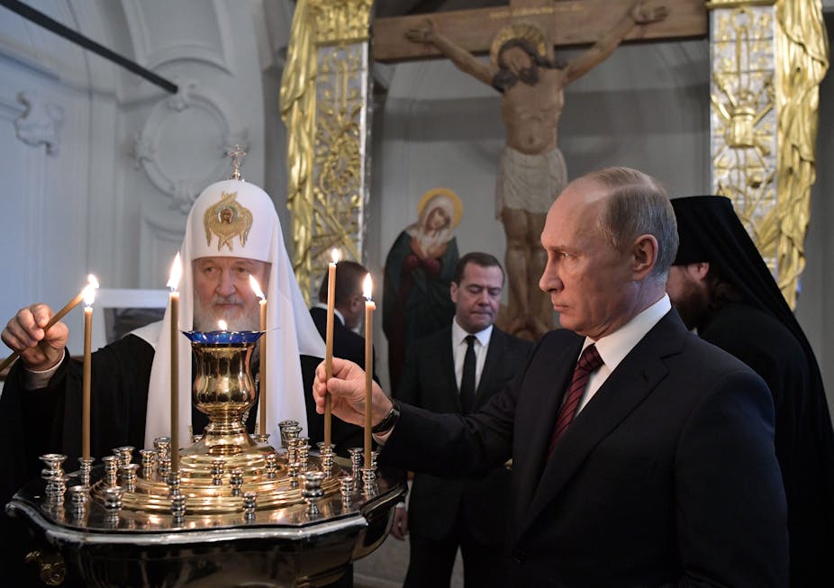 Patriarch of Russia Kirill and Vladimir Putin lighting candles at a monastery with an image of Christ in the background.
