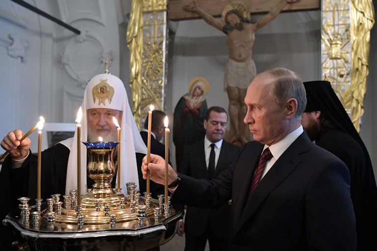 Patriarch of Russia Kirill and Vladimir Putin lighting candles at a monastery with an image of Christ in the background.
