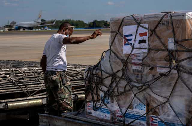 A man points, a pallette is in front of him with boxes and rope and an image of the Cuban and Syrian flag