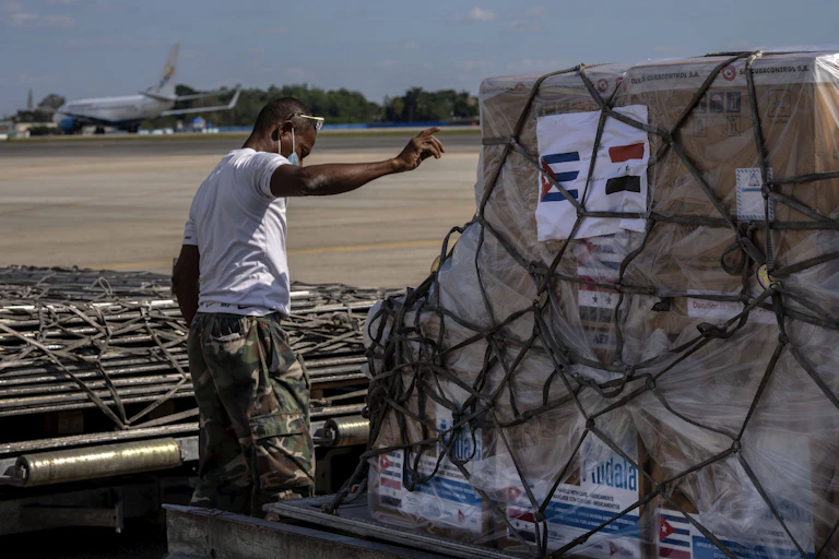 A man points, a pallette is in front of him with boxes and rope and an image of the Cuban and Syrian flag