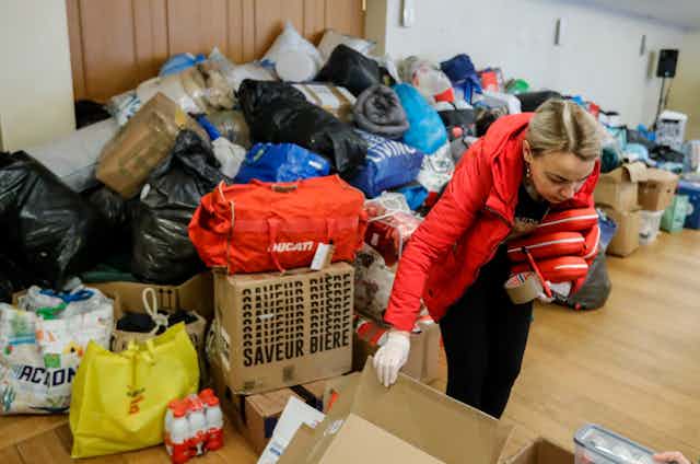 A woman picks up a cardboard box, behind her is a massive pile of bags and boxes filled with items