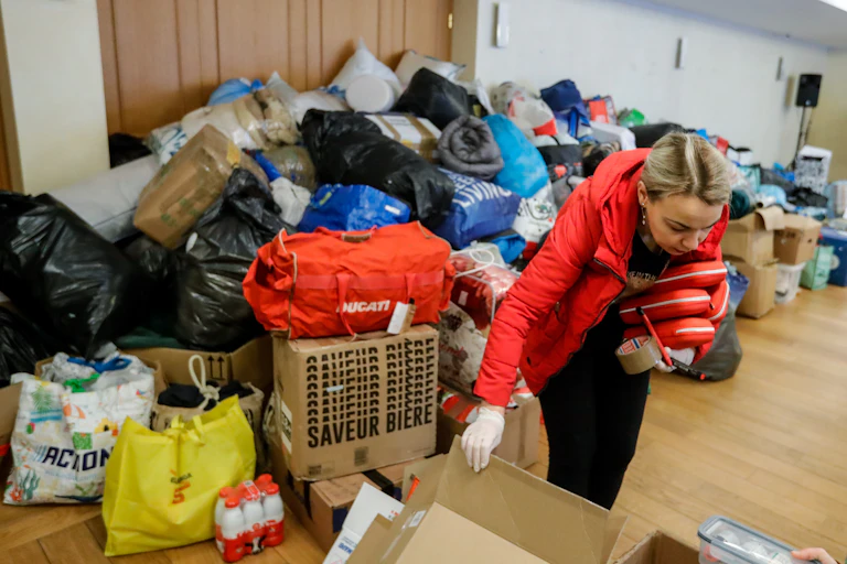A woman picks up a cardboard box, behind her is a massive pile of bags and boxes filled with items