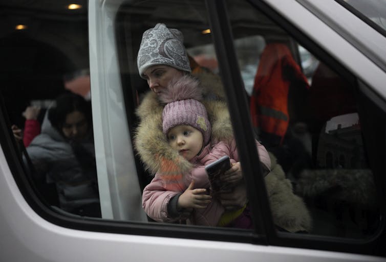 A woman holding a child, both dressed in winter coats and hats, as seen through a train window