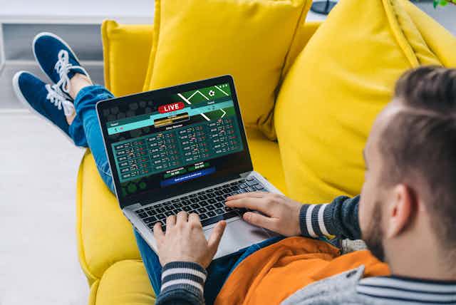 A man sits on a couch with a laptop, on which a sports betting programme is open.
