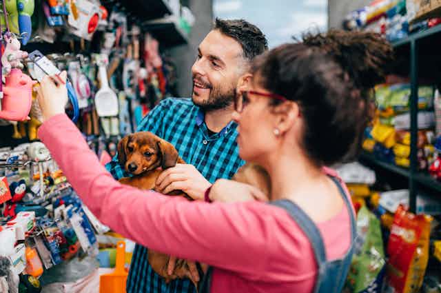 A smiling couple holds a small brown dachshund while browsing dog toys inside a shop