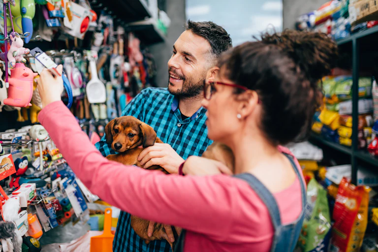 A smiling couple holds a small brown dachshund while browsing dog toys inside a shop