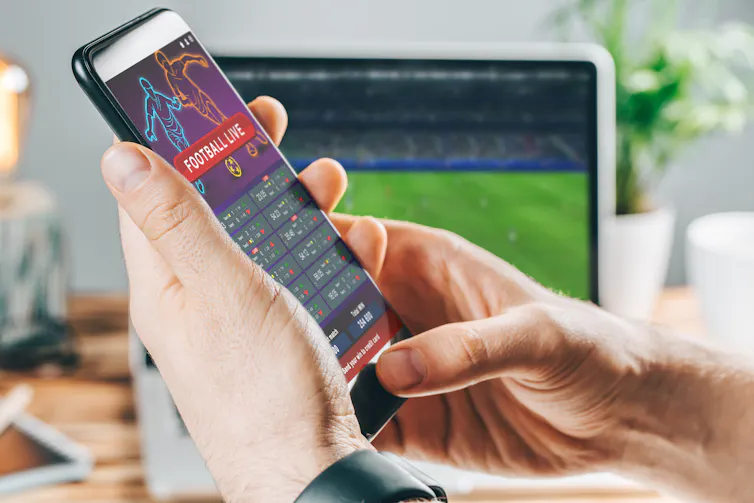 A hand holds a smartphone with a sports betting app open, in front of a laptop screen displaying a sporting match.