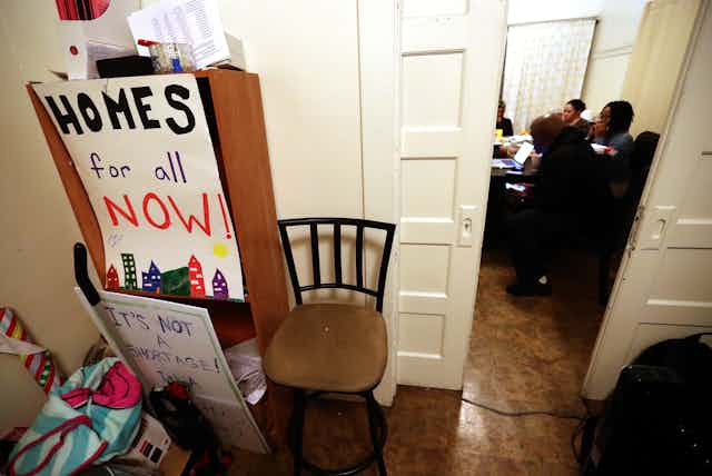A sign reading "Homes for all now" sits in front of an open door revealing people inside a room seated around a table.