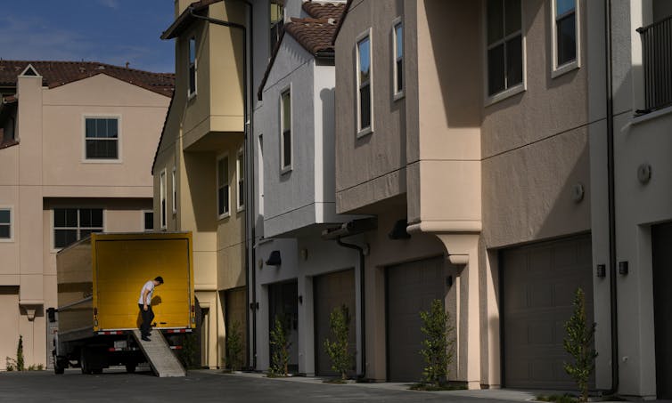 A man is shown outside of a moving truck, next to a row of new attached houses.