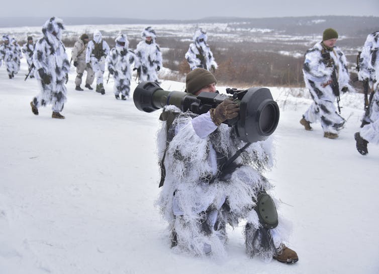 a soldier wearing a sniper's white winter camoflauge suit holds an anti-tank missile while other soldiers walk behind him on snowy hills.