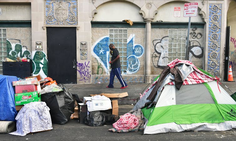 A man walks past a building with graffiti, in front of tents and boxes.