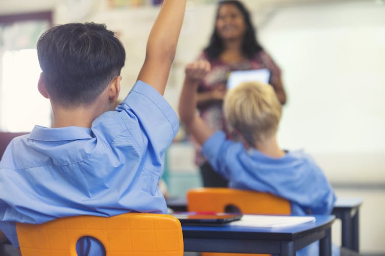 A classroom with young students, with their hands raised.