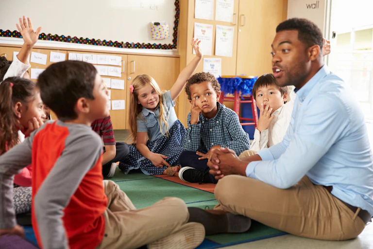 A male teacher sits crosslegged on a school carpet with a circle of primary-school children holding up their hands to answer.