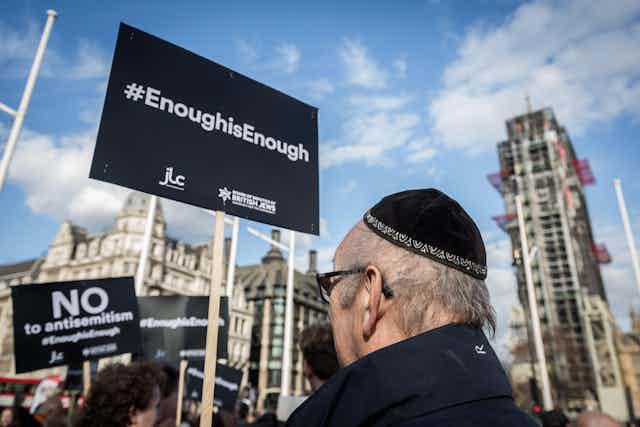 A man in a black yarmulke stands in the street surrounded by protest placards.