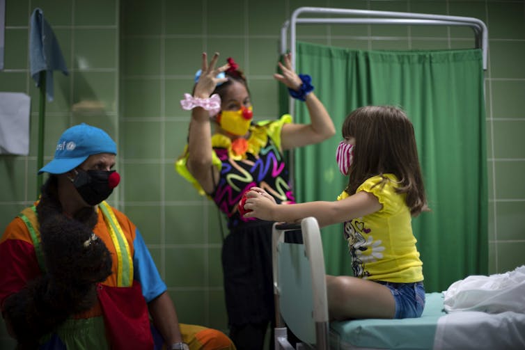 A girl sits on a hospital bed, wearing a face mask with her legs crossed. A clown wearing yellow entertains her.
