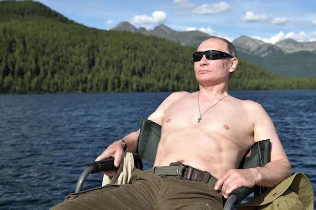 A man sitting next to a lake, shirtless and wearing sunglasses.