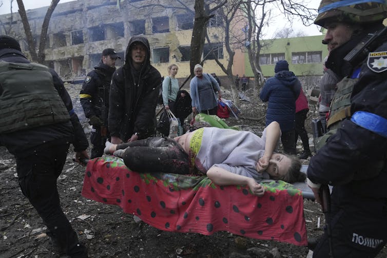 a pregnant woman lies on a stretcher as volunteer workers carry her over rubble with a destroyed building in the background