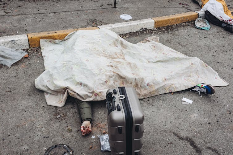 A dead body lying under a bloody white cloth, with a suitcase next to it.