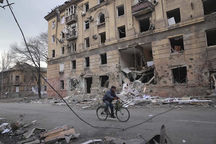 A man rides a bicycle passed a destroyed apartment building.