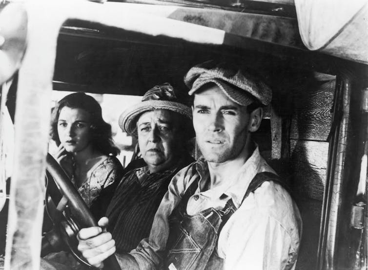 Two women and a man in old clothing look out from a car window in a black and white photograph.